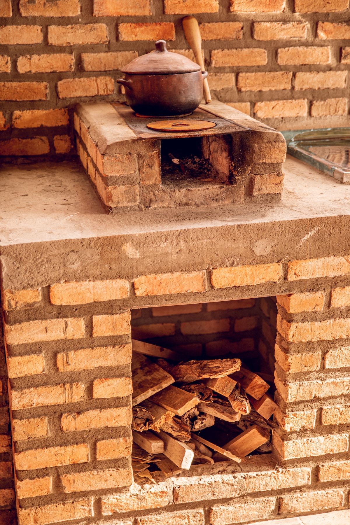 A rustic brick stove is depicted in the kitchen area, featuring a pot resting on top. The lower section reveals a compartment filled with neatly stacked firewood, enhancing the space's functionality.