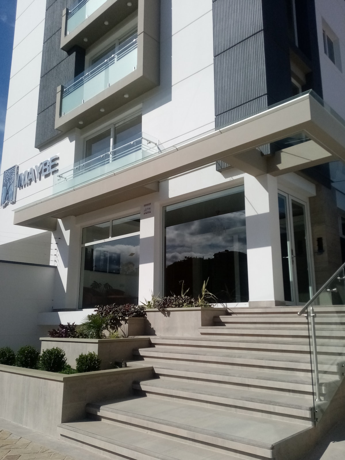 The exterior of a modern building is presented, featuring a clean design with large glass windows and a stylish entrance. Steps lead up to the entrance, surrounded by neatly arranged plants and landscaping, with a clear blue sky visible above.