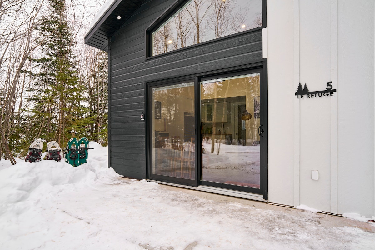 A modern building entrance is framed by snow, highlighting the large glass doors that offer a view into the interior. Two snowmobiles are parked nearby, surrounded by tall trees that enhance the serene winter setting.