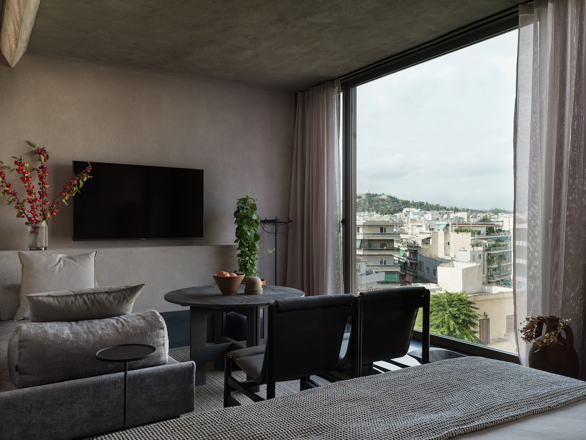 A modern living area is showcased with a large window offering views of the city. A small round table is surrounded by black chairs, while a plush gray sofa and decorative plants enhance the space. A wall-mounted television is positioned for easy viewing.