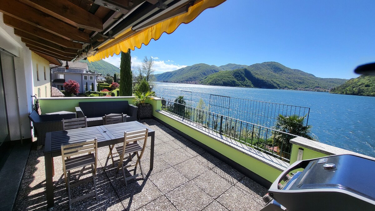 A spacious terrace is seen with a dining area featuring a large table surrounded by wooden chairs. A grill is placed nearby, while the stunning view of the lake and mountains is visible in the background, complemented by greenery along the edge.