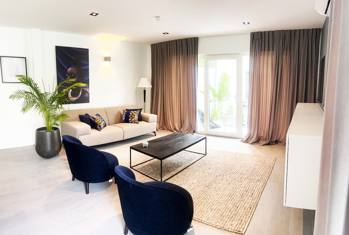 A modern living room is depicted with a light-colored sofa adorned with patterned cushions. Two navy blue chairs complement a sleek black coffee table atop a textured area rug. Large windows with floor-length curtains allow natural light to illuminate the space.