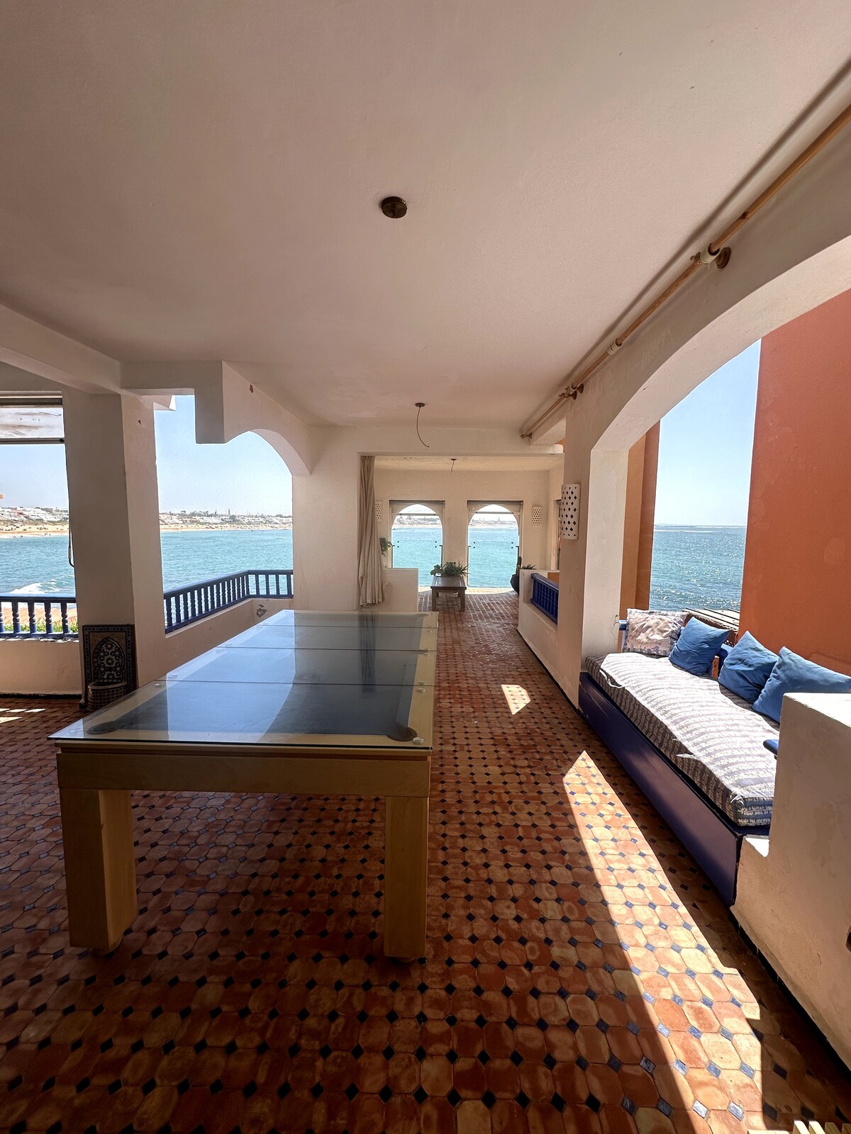 A spacious terrace is visible, featuring a large glass dining table and cushioned seating along the railing. Sunlight floods the area, highlighting the tiled floor and the nearby view of the water through arched openings.