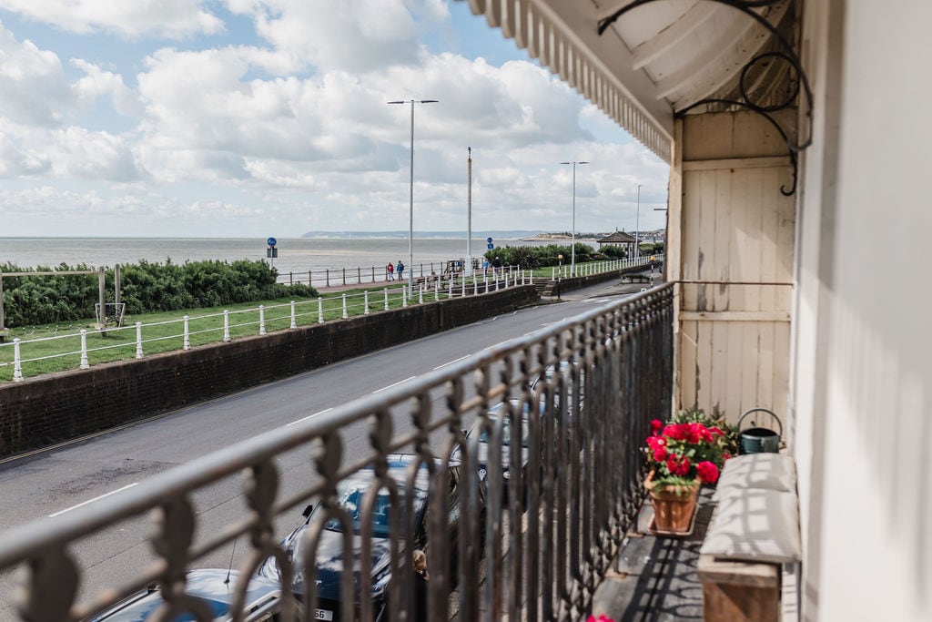 A charming balcony offers a view of the ocean beyond the street. Decorative planters with bright flowers are placed on the ledge, while a wrought iron railing adds character. The scene captures the picturesque coastline under a partly cloudy sky.