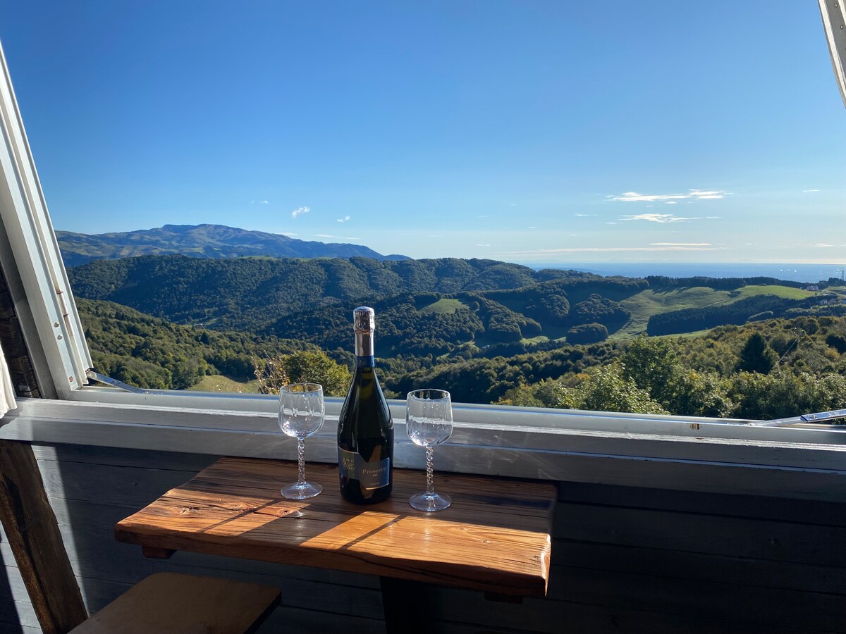 A wooden table is set with two elegant glasses and a bottle of sparkling wine, positioned in front of an open window. A sweeping view of green hills and valleys stretches into the distance under a clear blue sky.