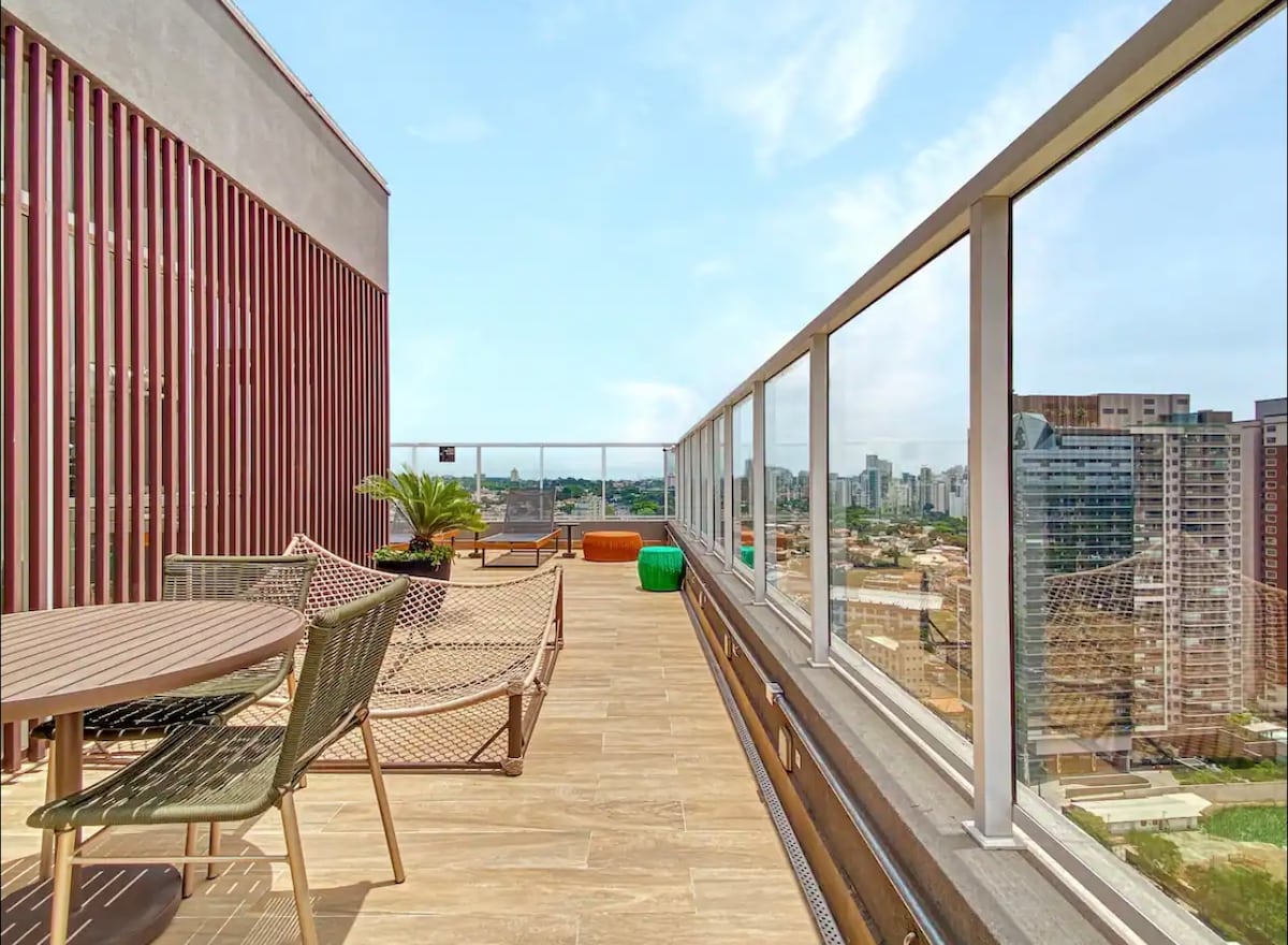 An expansive balcony stretches across the unit, featuring a wooden table surrounded by chairs. Comfortable lounge seating is visible alongside potted plants. A panoramic view of São Paulo's skyline is highlighted beyond the glass railing, creating an inviting outdoor space for relaxation.