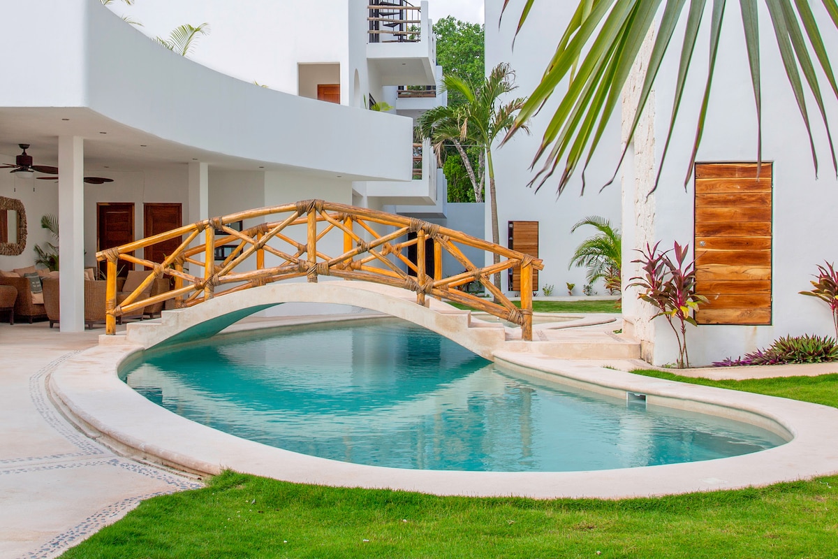 An elegant private plunge pool is framed by manicured green grass and surrounded by lush tropical plants. A wooden bridge arches gracefully over the water, connecting different areas of the property, while white walls and natural textures enhance the serene environment.
