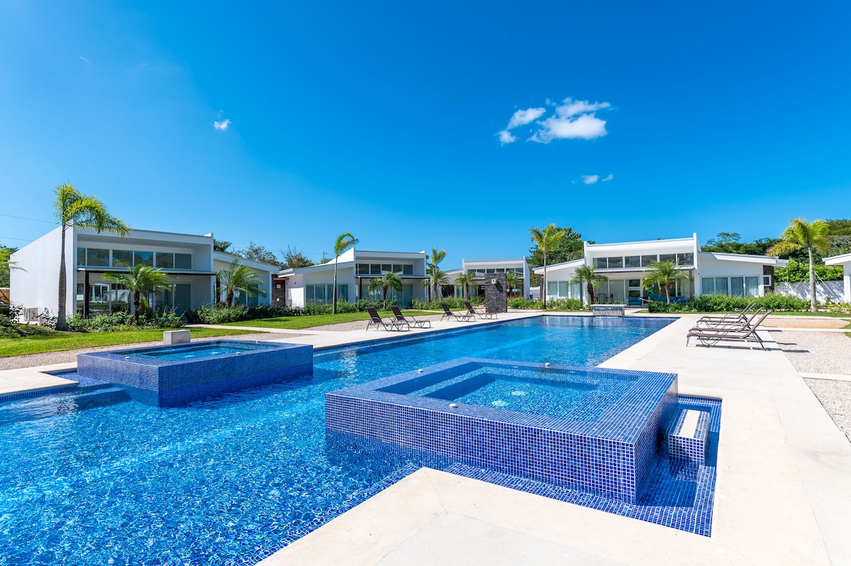 A clear blue sky overlooks a shared salt-water pool, featuring vibrant blue tiles and jetted tubs. Surrounding palm trees and lounge chairs frame the area, while modern villa structures provide a serene backdrop, showcasing the tranquil ambiance of this gated community.