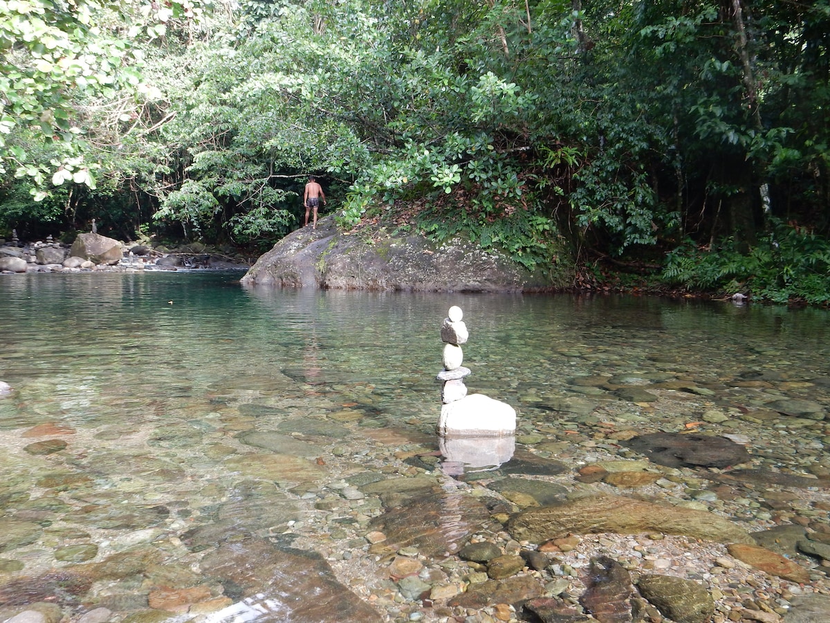 A tranquil swimming area in a forest setting is depicted, featuring clear water over smooth stones. A stack of stones stands prominently in the foreground, while lush greenery surrounds the area. A person is seen in the background, near a large rock.