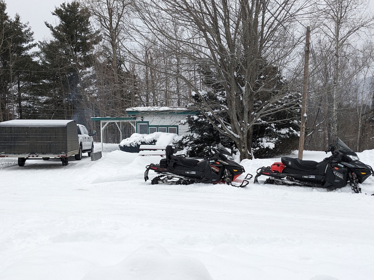 A snowy outdoor scene features two parked snowmobiles on a snow-covered driveway, with trees in the background. A vehicle with a trailer is positioned nearby, and a cabin is visible behind the trees, surrounded by snow-laden ground.