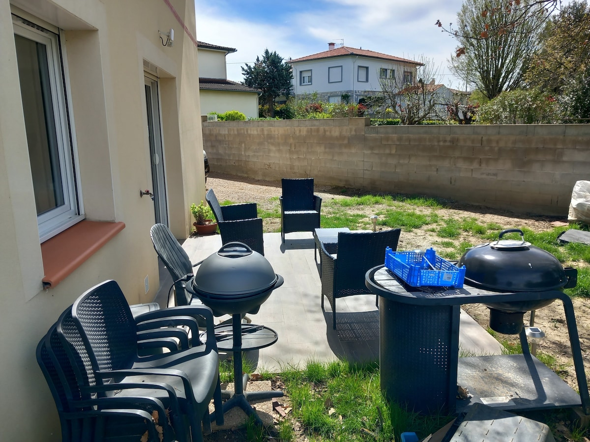An outdoor area features several black patio chairs arranged around a small table, alongside a charcoal barbecue grill. A blue basket sits on a table, and the green grass adds a natural element to the space. A wall provides privacy from the surroundings.
