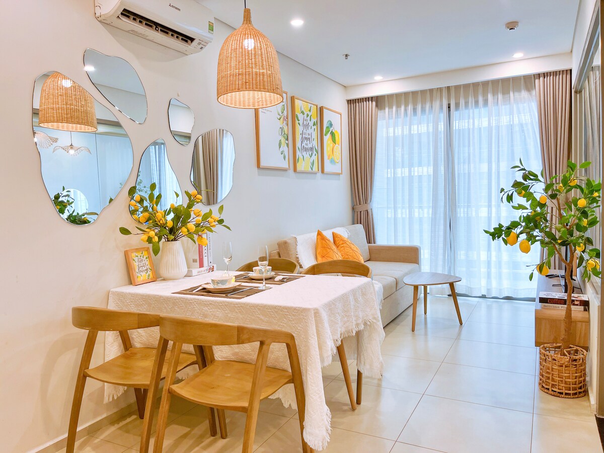 A well-lit dining area features a round table surrounded by two wooden chairs, complemented by a soft white tablecloth. A cozy sofa is situated nearby, and decorative mirrors reflect light. Framed artwork and a potted plant add a touch of color to the space.