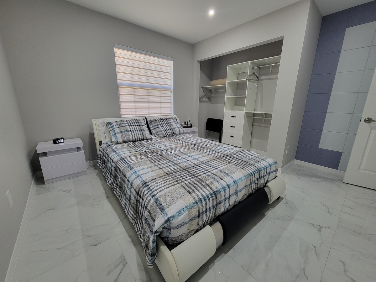 A spacious sleeping area features a queen-sized bed with a plaid blanket. A bedside table holds a clock, and a closet with shelves is visible. Light enters through a window covered by light blinds, highlighting the neutral wall tones and tiled floor.