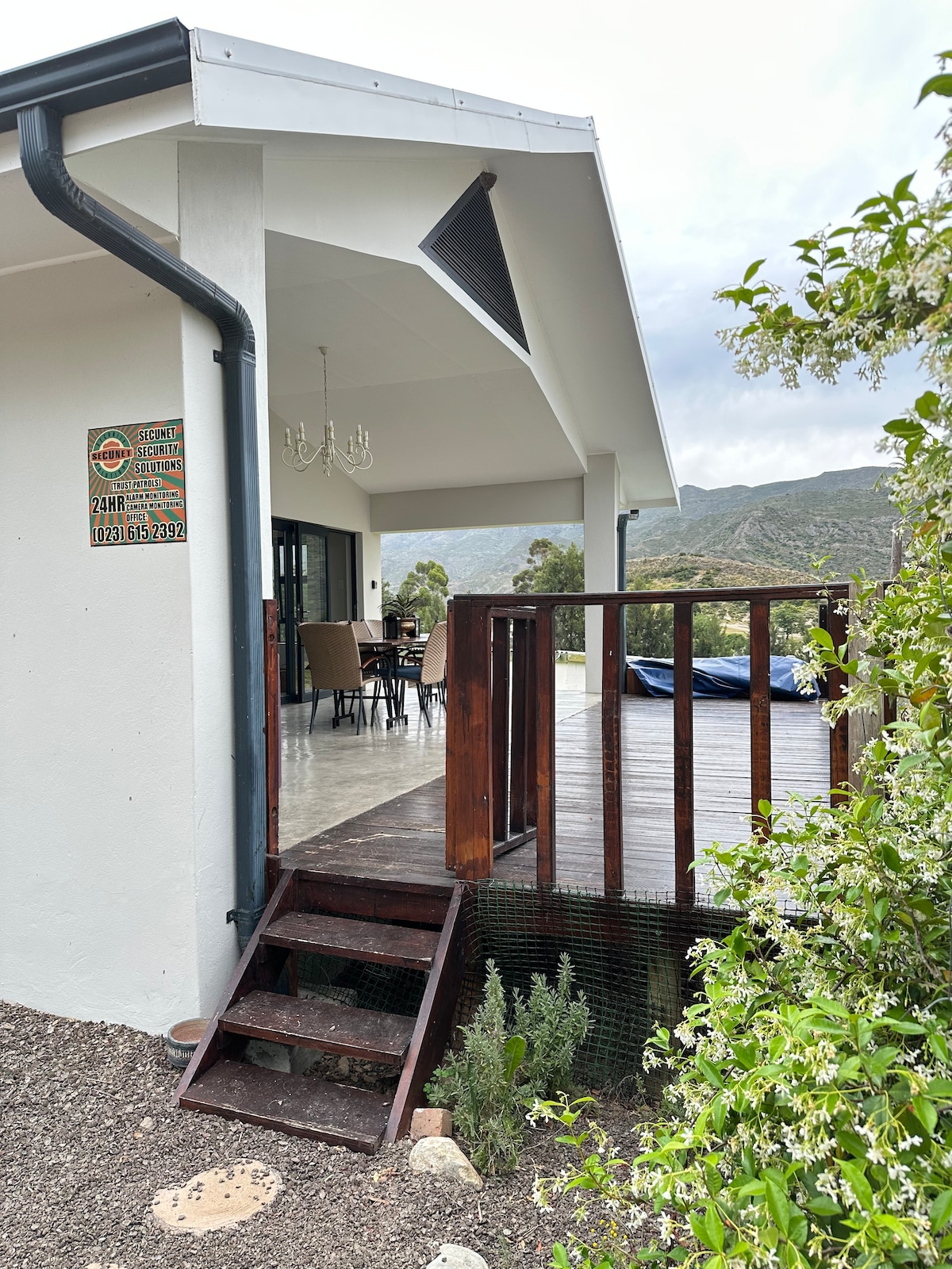 The exterior features a welcoming porch with steps leading up to a covered area. A wooden railing surrounds the deck, and a dining set is visible in the background. Lush greenery and a mountain view enhance the serene setting.