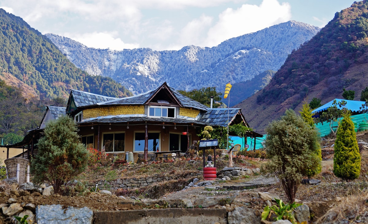 The exterior of a charming structure is visible, framed by mountains in the background. A stone pathway leads to the entrance, while shrubs and small trees provide greenery. The roof features wooden accents and a mixture of materials, complementing the natural landscape.