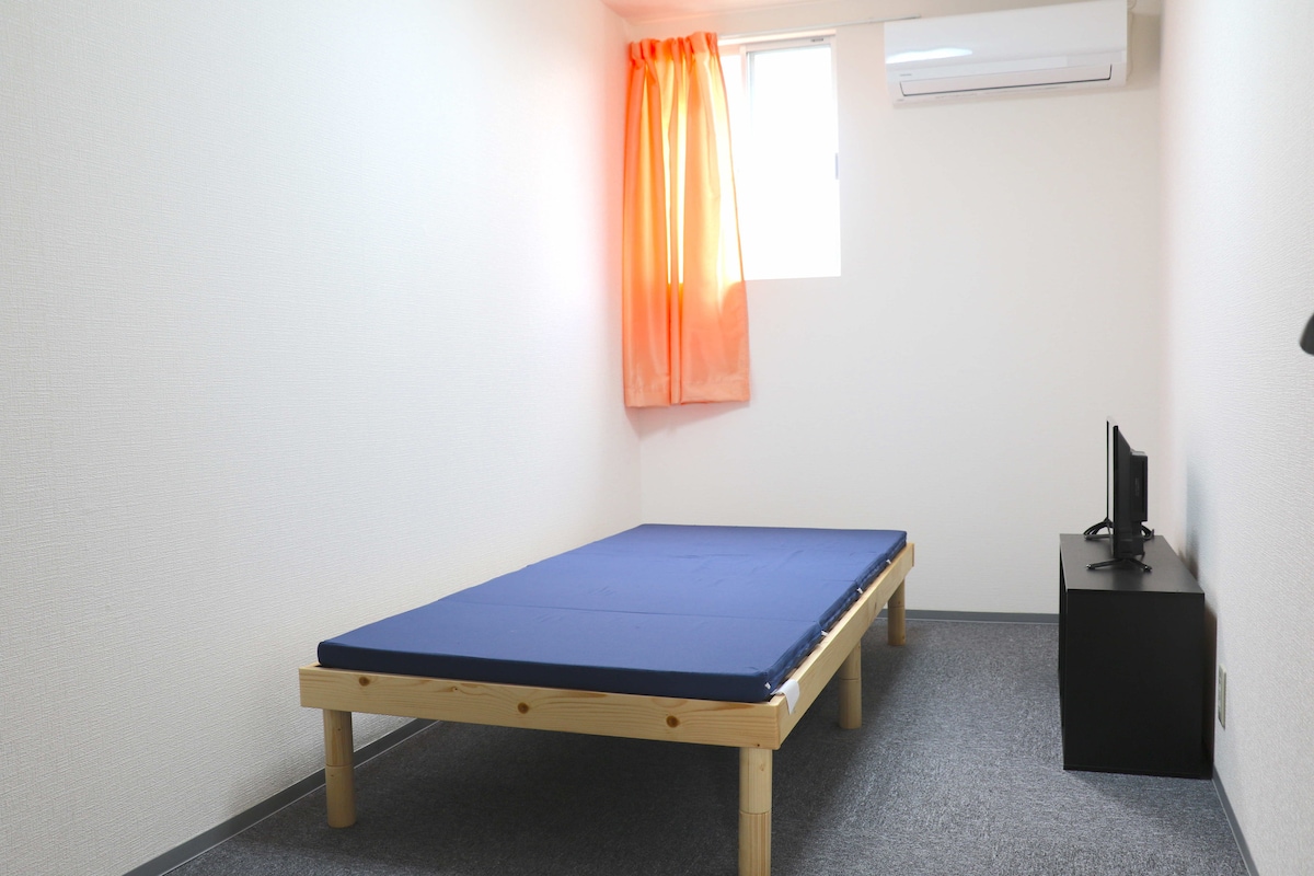 A simple sleeping area features a low wooden bed frame with a blue mattress. Natural light enters through a window dressed with an orange curtain. A small television is positioned on a black stand against the wall, complementing the minimalistic design.