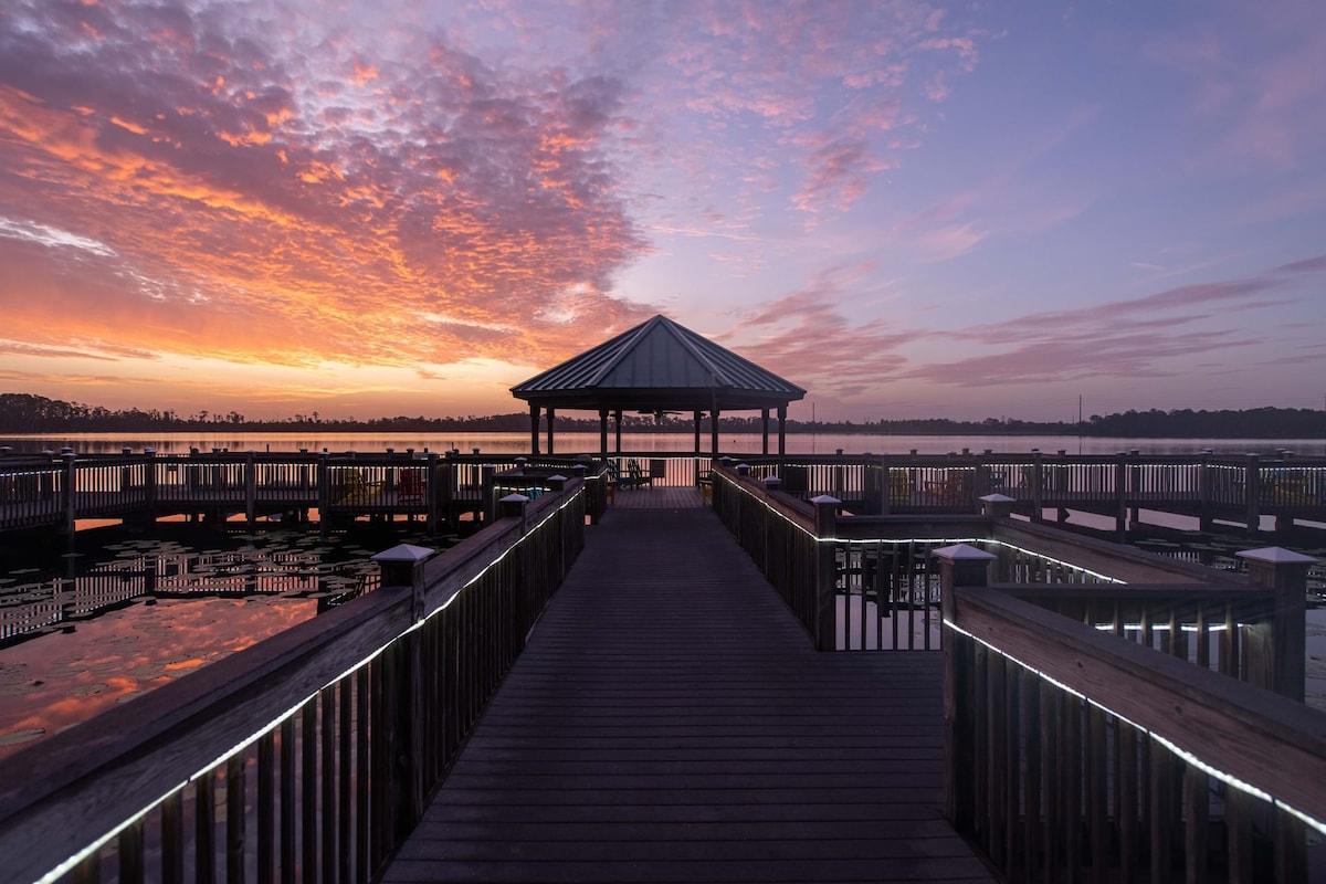 A wooden pier leads to a gazebo, surrounded by calm water reflecting a vibrant sunset. The sky is painted with soft hues of orange and pink, creating a serene atmosphere for relaxation. The walkway is lined with subtle lighting for visibility.