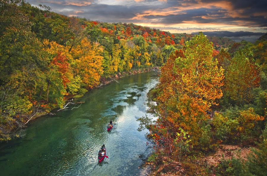 A tranquil river winds through a vibrant landscape, showcasing autumn foliage in shades of orange, yellow, and green. Two canoes glide along the water, reflecting the colorful trees lining the banks under a dramatic sky.