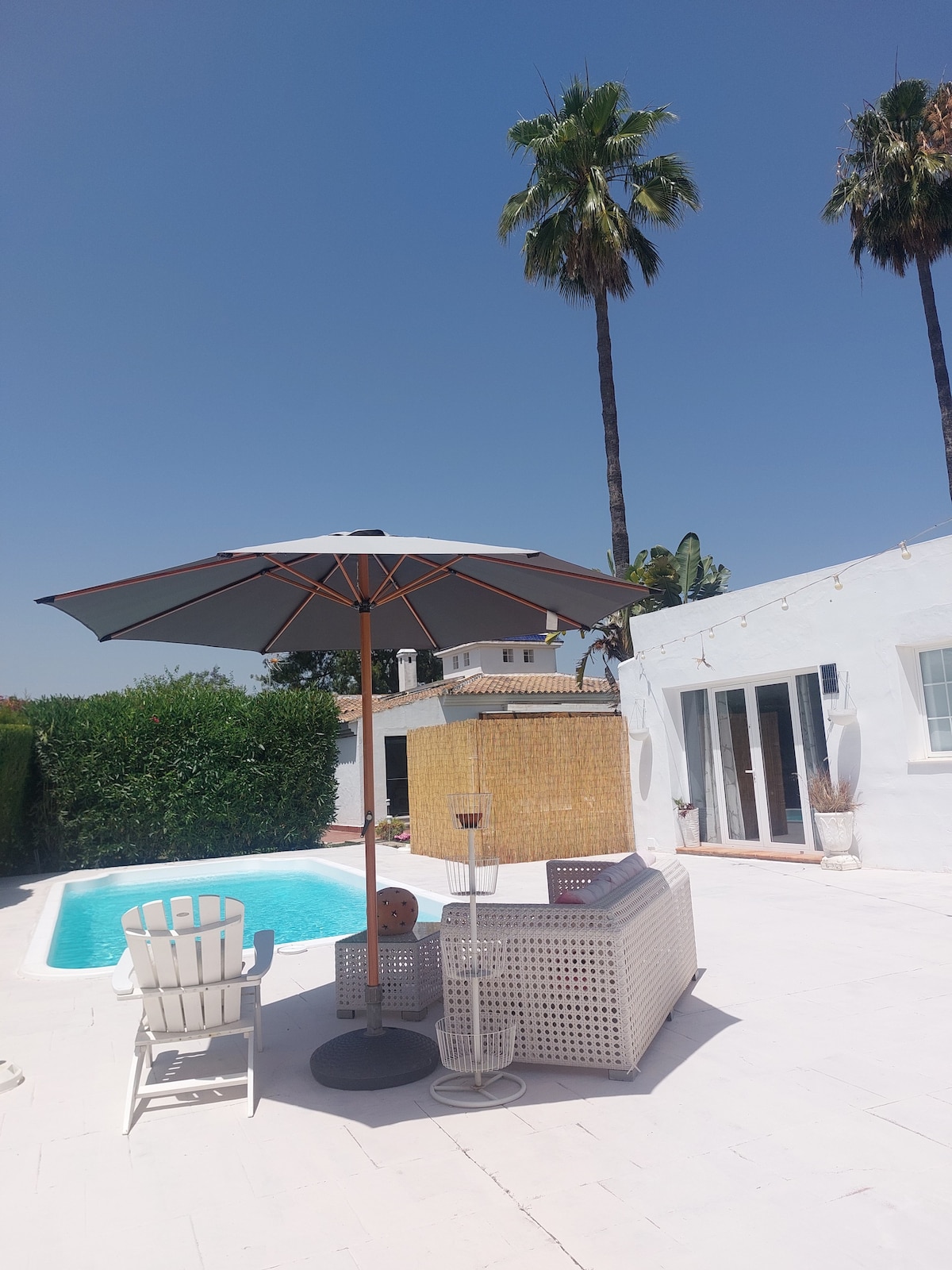 A private outdoor area features a shimmering pool surrounded by a white deck. A large umbrella provides shade over a stylish seating arrangement, which includes a chair and a small table. Tall palm trees and lush greenery create a tropical atmosphere.
