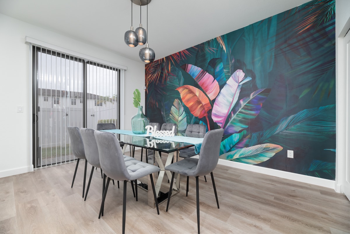 A modern dining area is highlighted by a glass table surrounded by six textured grey chairs. Behind the table, a vibrant tropical mural adds a splash of color, while natural light filters in through large sliding doors, showcasing the outdoor space.