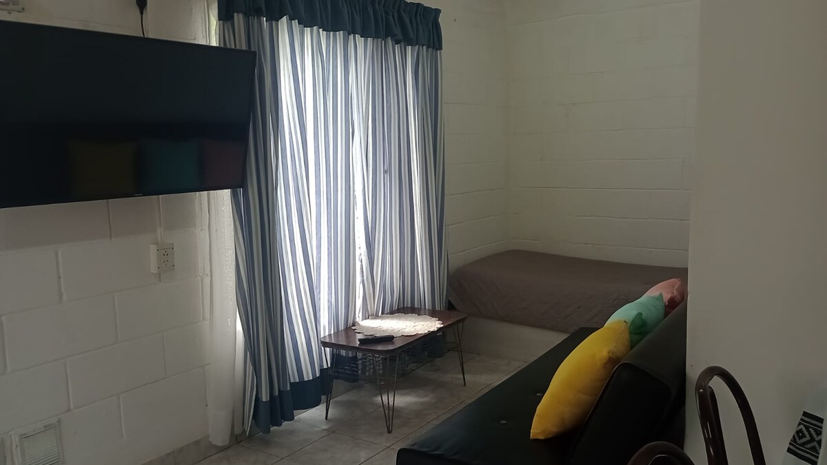 A living space is presented with a large flat-screen TV mounted on a white wall. Natural light enters through tall, striped curtains, highlighting a simple bed against the wall and a dark sofa in the foreground. A low coffee table is placed in the center.
