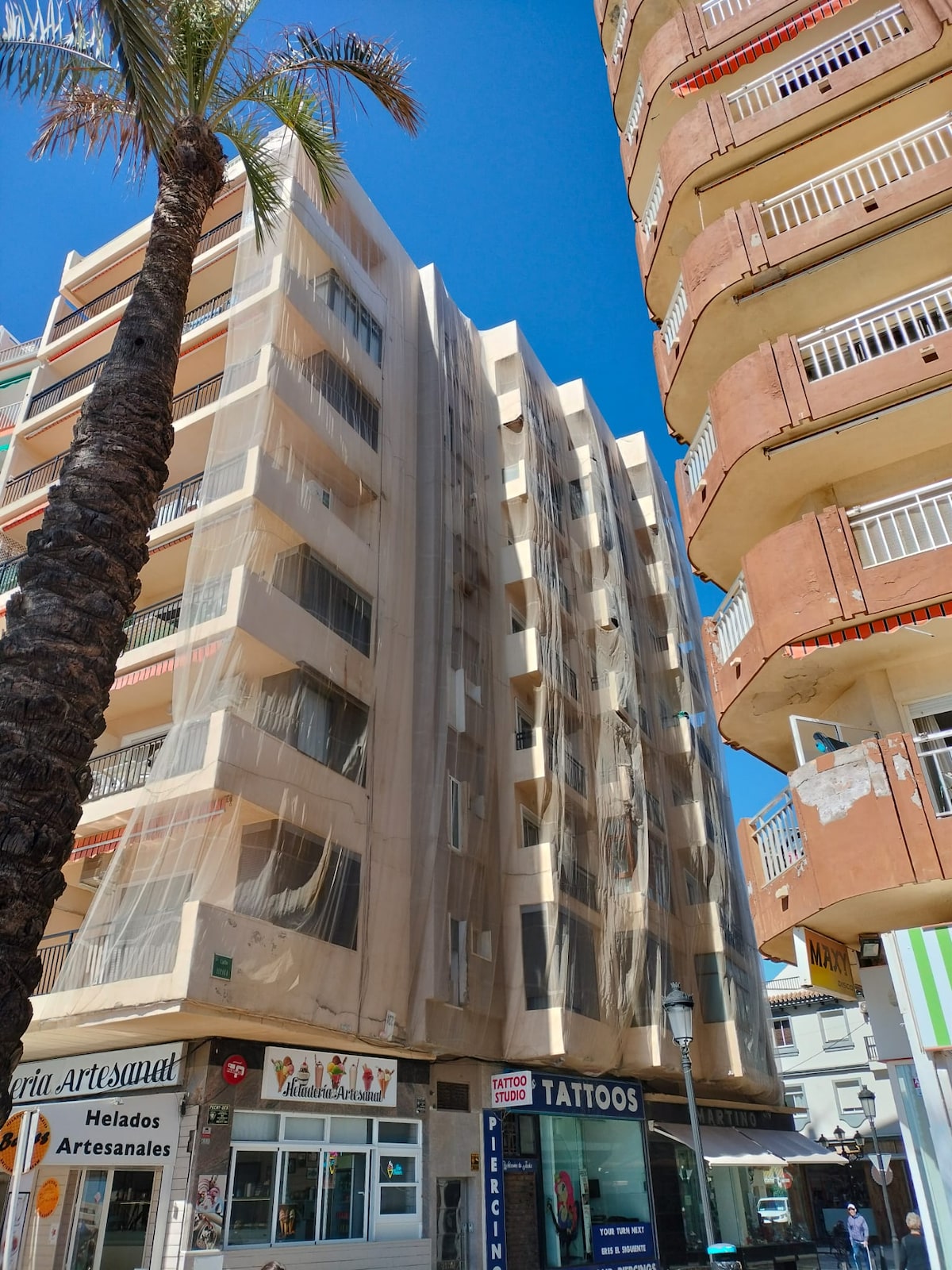 The exterior of a multi-story building is visible, covered in protective netting due to planned repairs. A prominent palm tree stands in the foreground, while adjacent buildings display various storefronts, contributing to the vibrant urban atmosphere.
