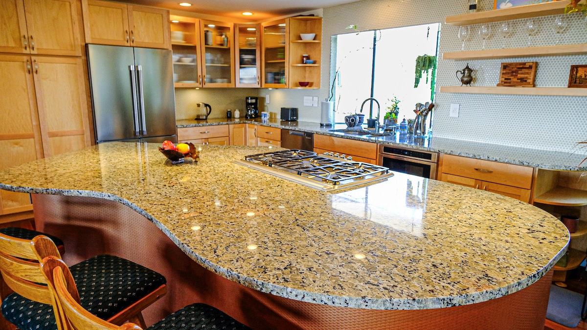 An inviting kitchen features an oversized granite countertop island with a curved design. Stainless steel appliances are present, along with wooden cabinetry and glass-front storage. A fruit bowl sits on the island, and natural light streams in from a window, illuminating the space.