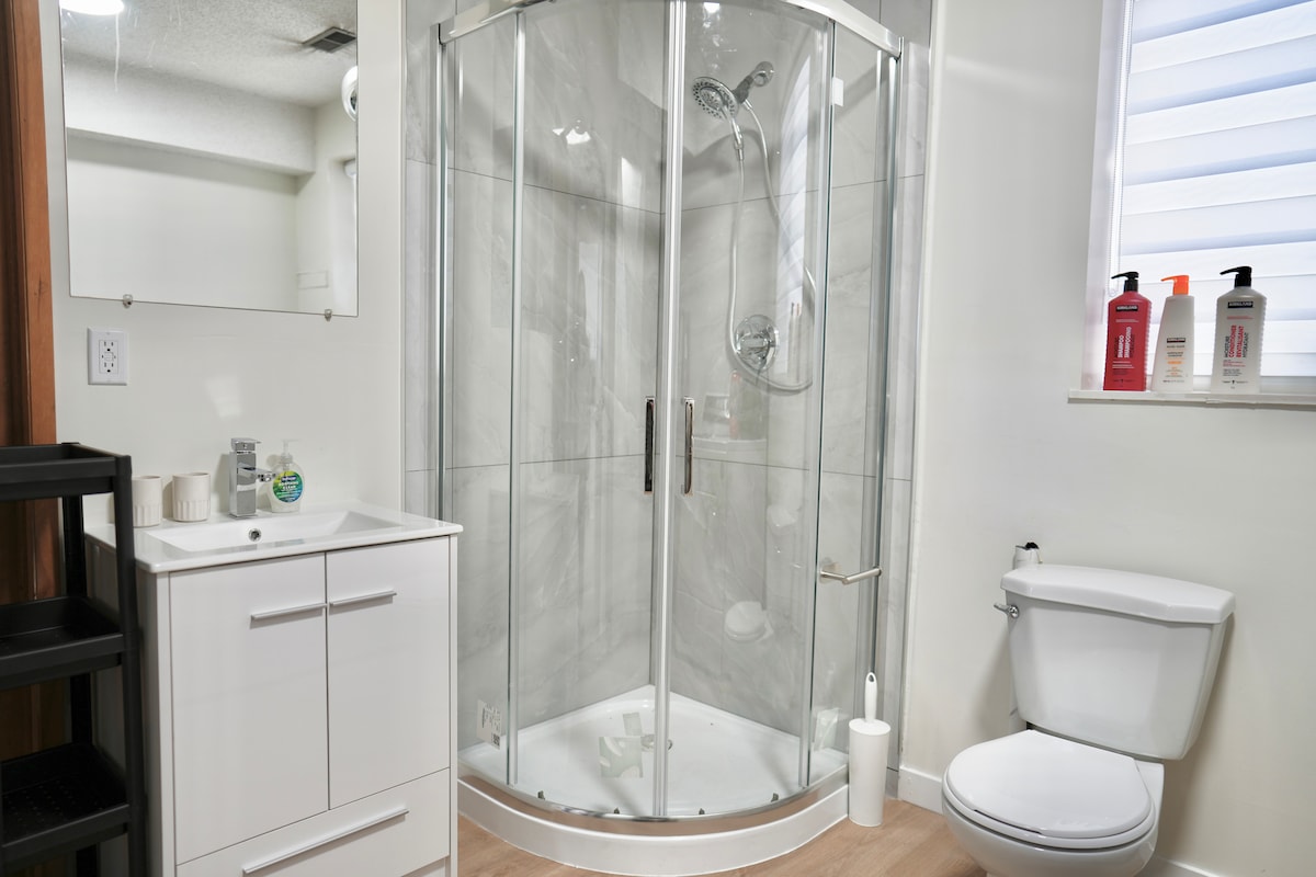 A modern bathroom is presented, featuring a curved glass shower with a rain showerhead. A sleek white vanity with toiletries is positioned beside the shower. A toilet and a small storage unit are situated in the corner, with light streaming in from a window above.