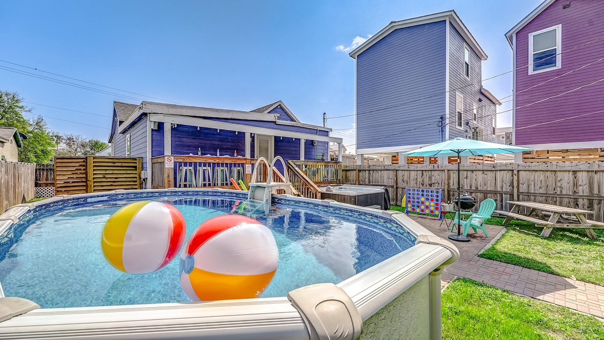 A vibrant backyard scene features a round pool with colorful beach balls floating. Nearby, a spacious patio area is visible, equipped with a table and sun umbrella. Lounge chairs are arranged around the pool, surrounded by grassy areas and paved pathways.