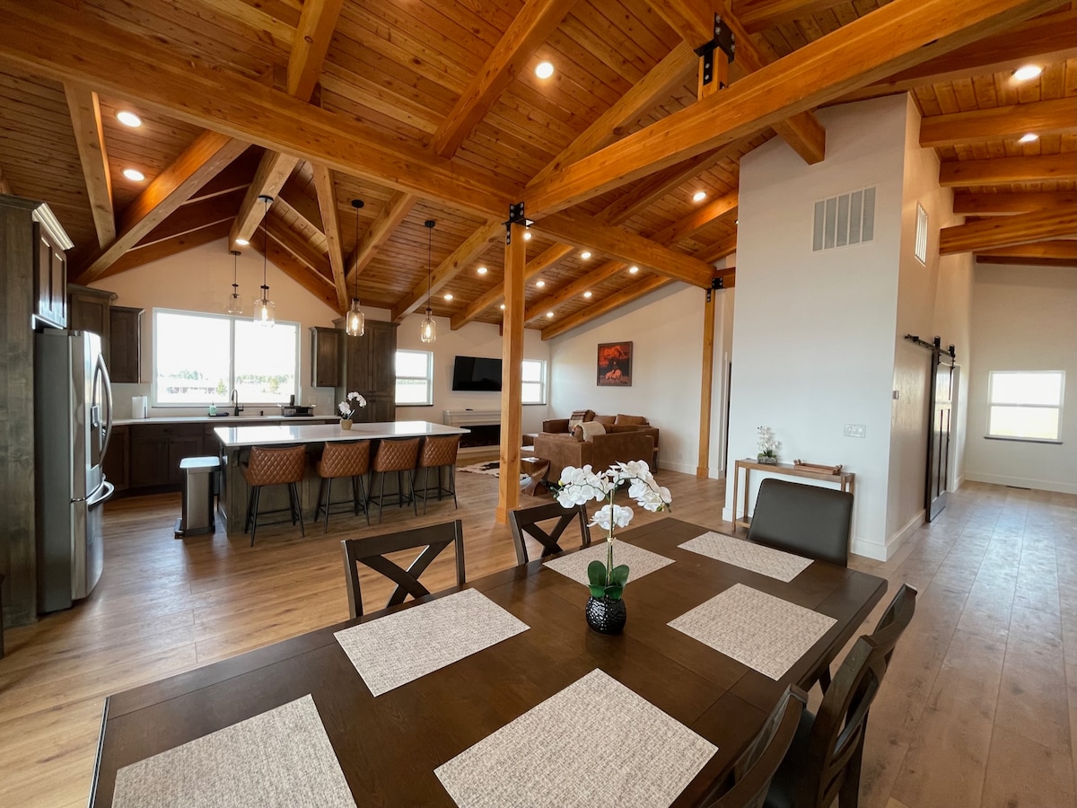 An open-concept living area features vaulted wooden ceilings and large windows, allowing natural light to fill the space. A dining table with place settings is situated near the kitchen and living area, which includes a comfortable seating arrangement and a modern entertainment setup.