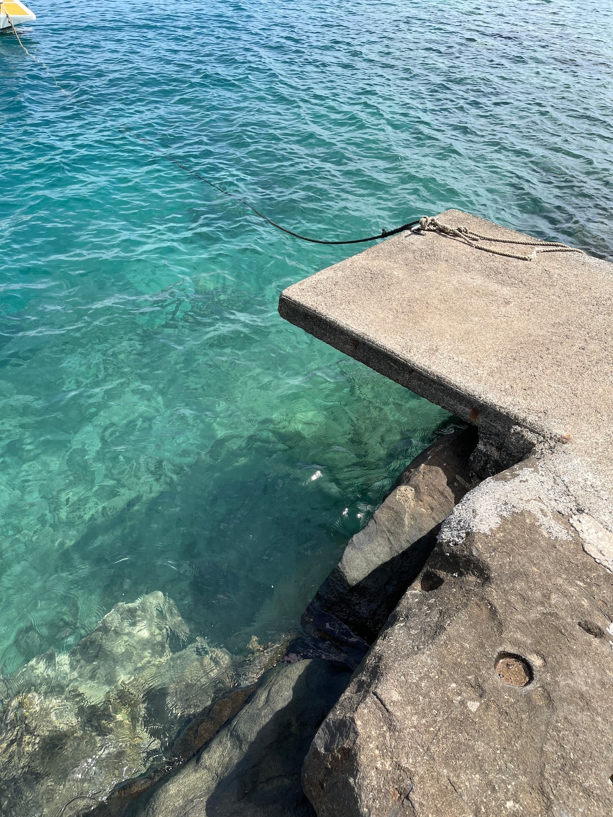 A concrete pier extends over calm, clear turquoise waters. Sunlight reflects off the surface, revealing a visible rocky seabed below. The image captures a section of the water just beside the edge of the pier, inviting an immersive experience of the marine environment.