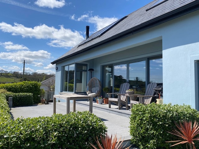 The exterior of the architect-designed house is visible, featuring a spacious patio with modern outdoor furniture. Lush greenery surrounds the area, and large glass doors open to the interior, inviting natural light. A clear blue sky enhances the serene atmosphere.