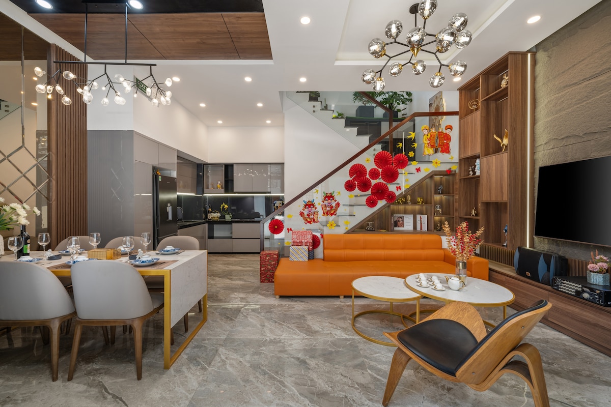 The open-concept living area features a stylish orange sofa paired with a wooden chair. A modern dining table can accommodate six guests. The kitchen area is seamlessly integrated, with sleek cabinetry and contemporary appliances. Decorative artwork adds a personal touch to the space.
