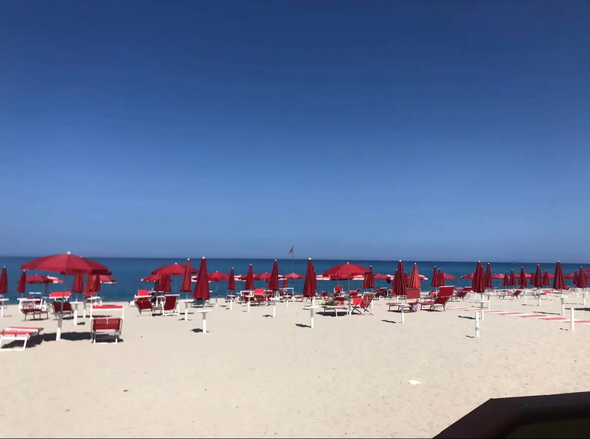The beach is lined with red umbrellas and lounge chairs, arranged neatly on the soft sand. The calm sea stretches in the background under a clear blue sky, creating a serene coastal setting.