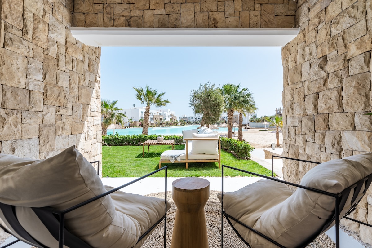 A stone archway frames a view of a lush garden and calm lagoon. Two comfortable lounge chairs with light cushions are positioned in the foreground, accompanied by a small wooden side table. Palm trees and sun loungers are visible in the background under a clear blue sky.