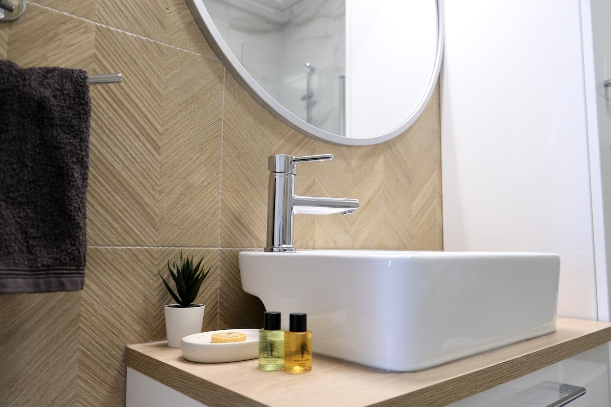 A modern bathroom countertop features a sleek, rectangular sink, complemented by a circular mirror above. A small plant and decorative items, including yellow and green bottled cosmetics, are arranged on the surface, alongside a textured gray towel hanging nearby.
