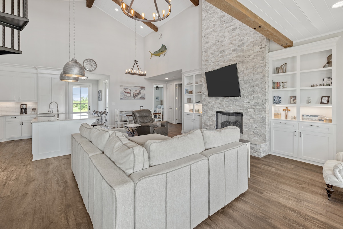 A spacious living area features a light-colored sectional sofa, offering ample seating. Natural light enters through large windows, illuminating the room. A stone fireplace serves as a focal point, while built-in shelves hold decorative items. The open layout connects to the kitchen and dining area.