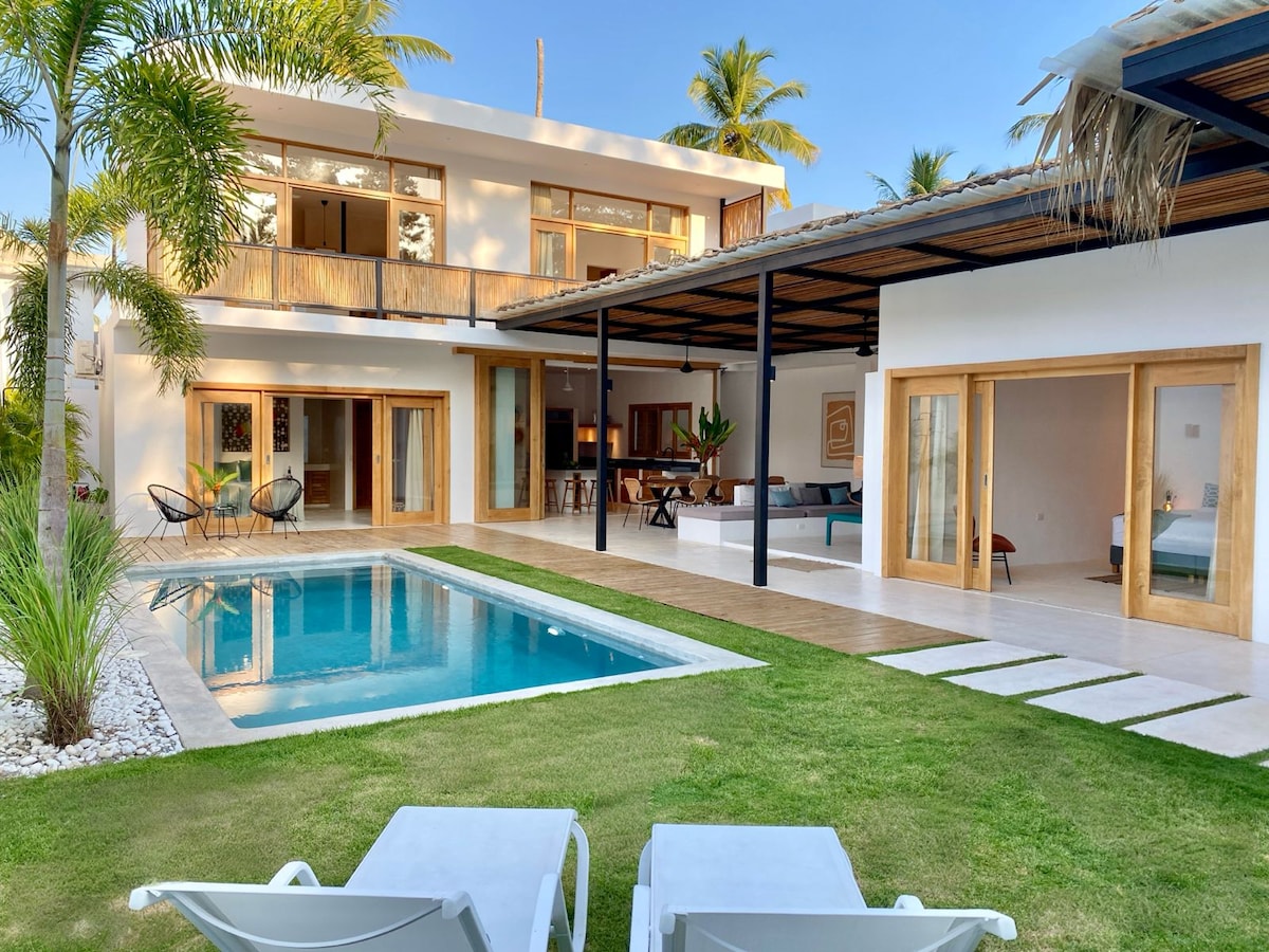 The exterior of the villa is showcased with a private pool, framed by a well-maintained lawn and palm trees. Large glass doors open up to the living area, creating a seamless flow between indoor and outdoor spaces. Lounge chairs are positioned near the poolside.