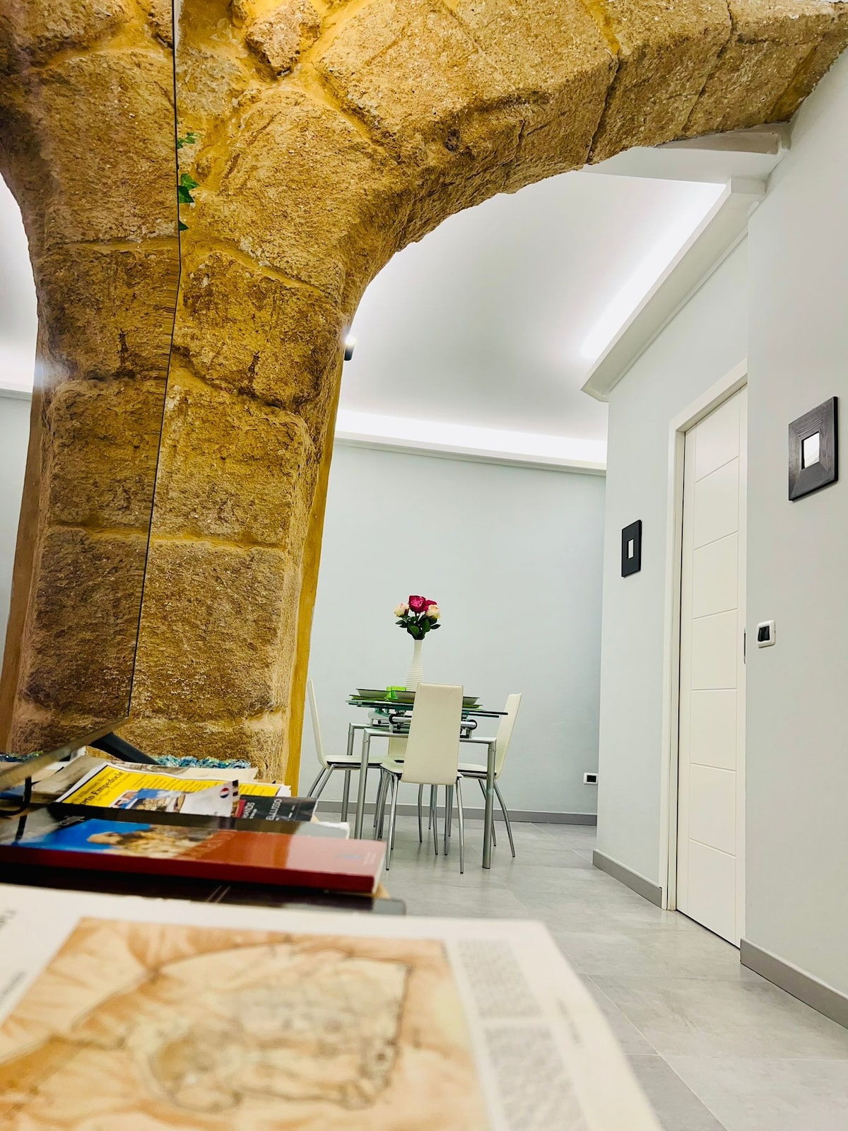 An interior view showcases a column of exposed stone, adding character to the space. A small dining table is set with a vase of flowers, and light-colored walls enhance the room's brightness. A doorway leads to additional areas of the apartment.