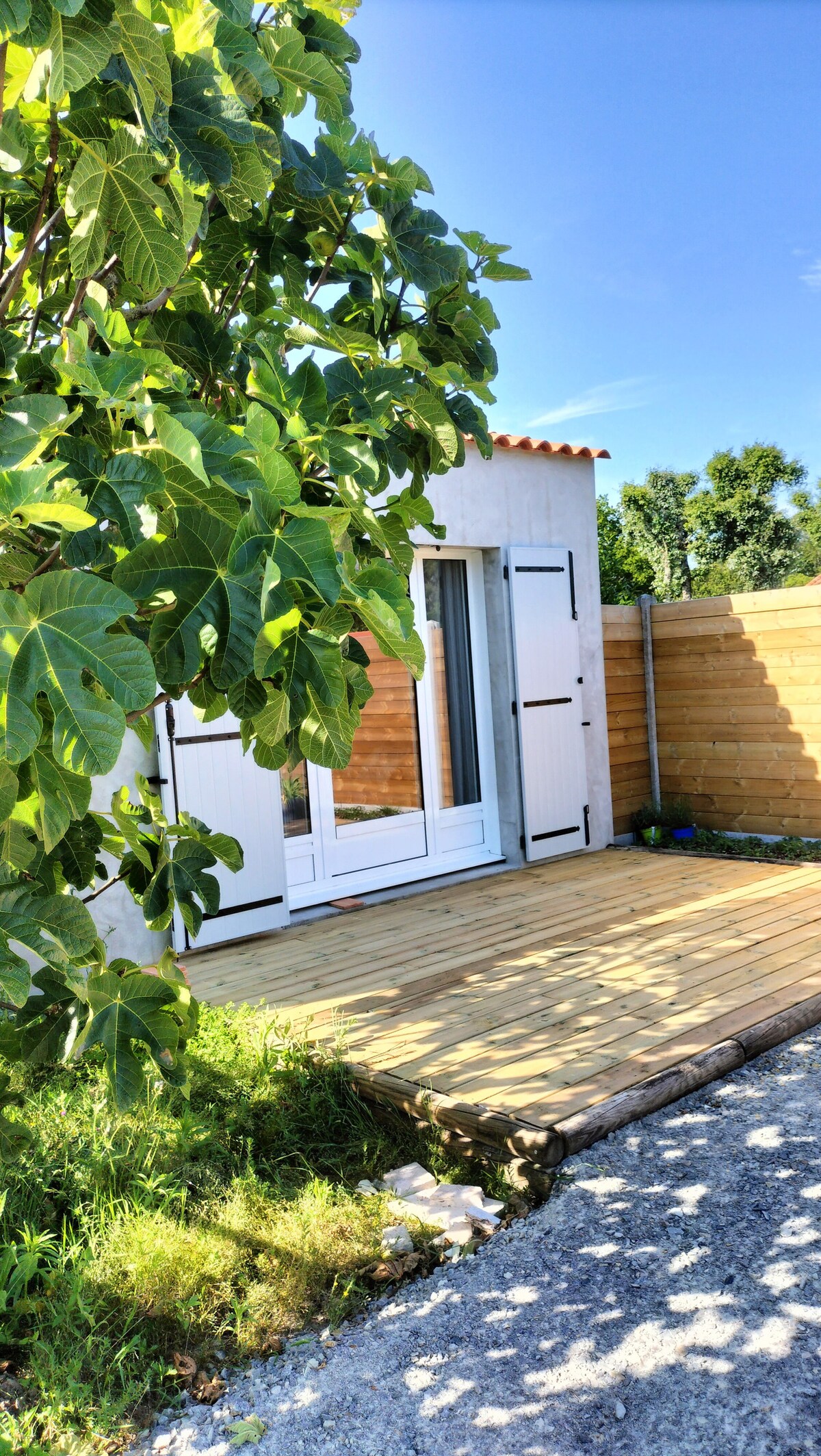 A serene outdoor area is depicted featuring a wooden deck in front of a neutral-colored exterior. Large, white-framed doors provide access, while lush green foliage from a nearby tree adds privacy and a natural touch to the space, complemented by a wooden fence.