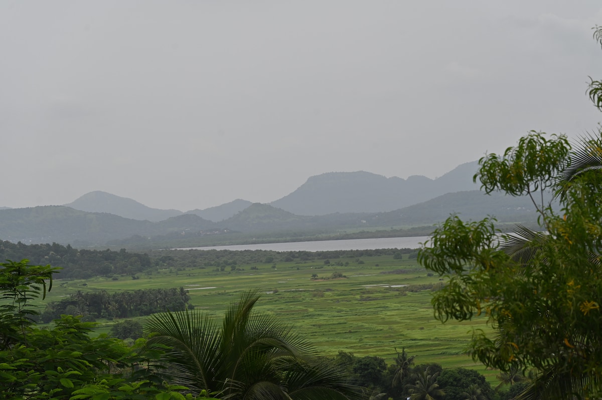 A serene landscape is captured, featuring rolling green hills and distant mountains under a cloudy sky. A calm body of water reflects the surrounding scenery, while lush foliage in the foreground adds depth to the natural view.