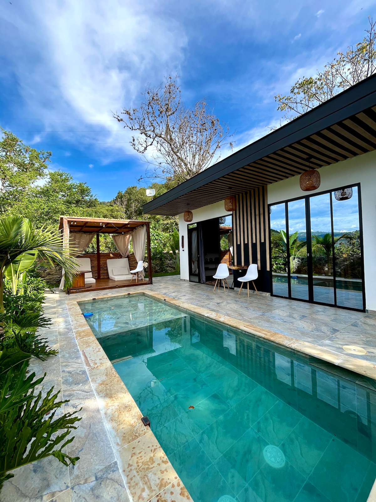 A modern home is surrounded by lush greenery, featuring a private swimming pool with clear water. Outdoor seating is available on a patio next to the pool, while expansive glass doors create a connection to the natural surroundings.