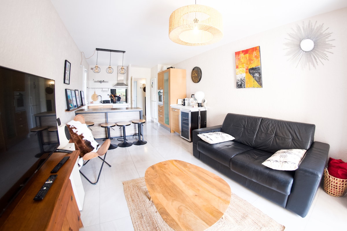 A living area features a black leather sofa positioned near a round wooden coffee table. A flat-screen television is mounted on the wall. In the background, an open kitchen is visible with modern appliances and bar seating provided by three stools. Artwork adorns the walls, adding a touch of design.
