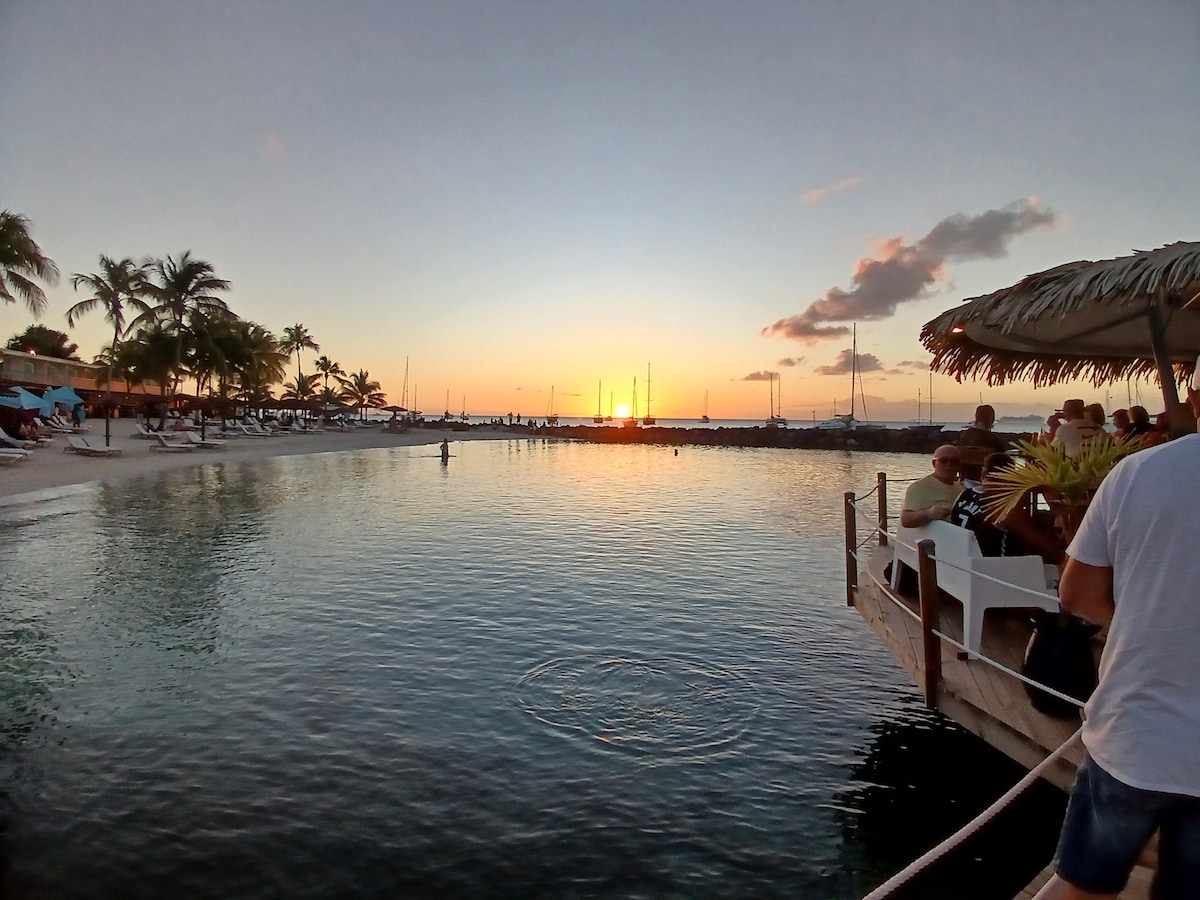 A serene seascape features calm waters reflecting a vibrant sunset. Palm trees frame the shoreline, and silhouettes of boats can be seen in the distance. The beach area is populated with lounge spaces, inviting relaxation as the day transitions into evening.