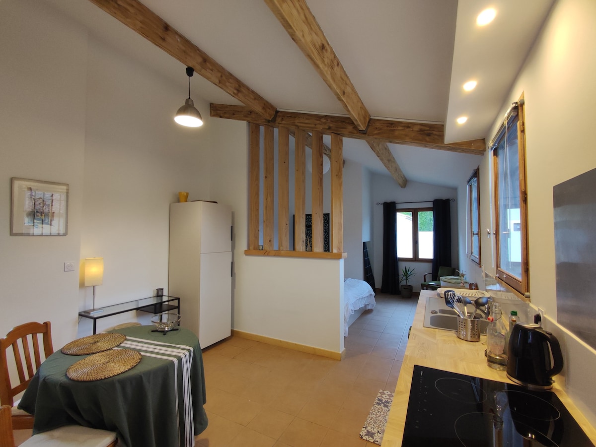 An open-plan studio features wooden beams overhead and a simple dining table set with placemats. A kitchenette with modern appliances is visible on one side, while a cozy sleeping area, partially separated, showcases a bed framed by natural light flowing through two windows.