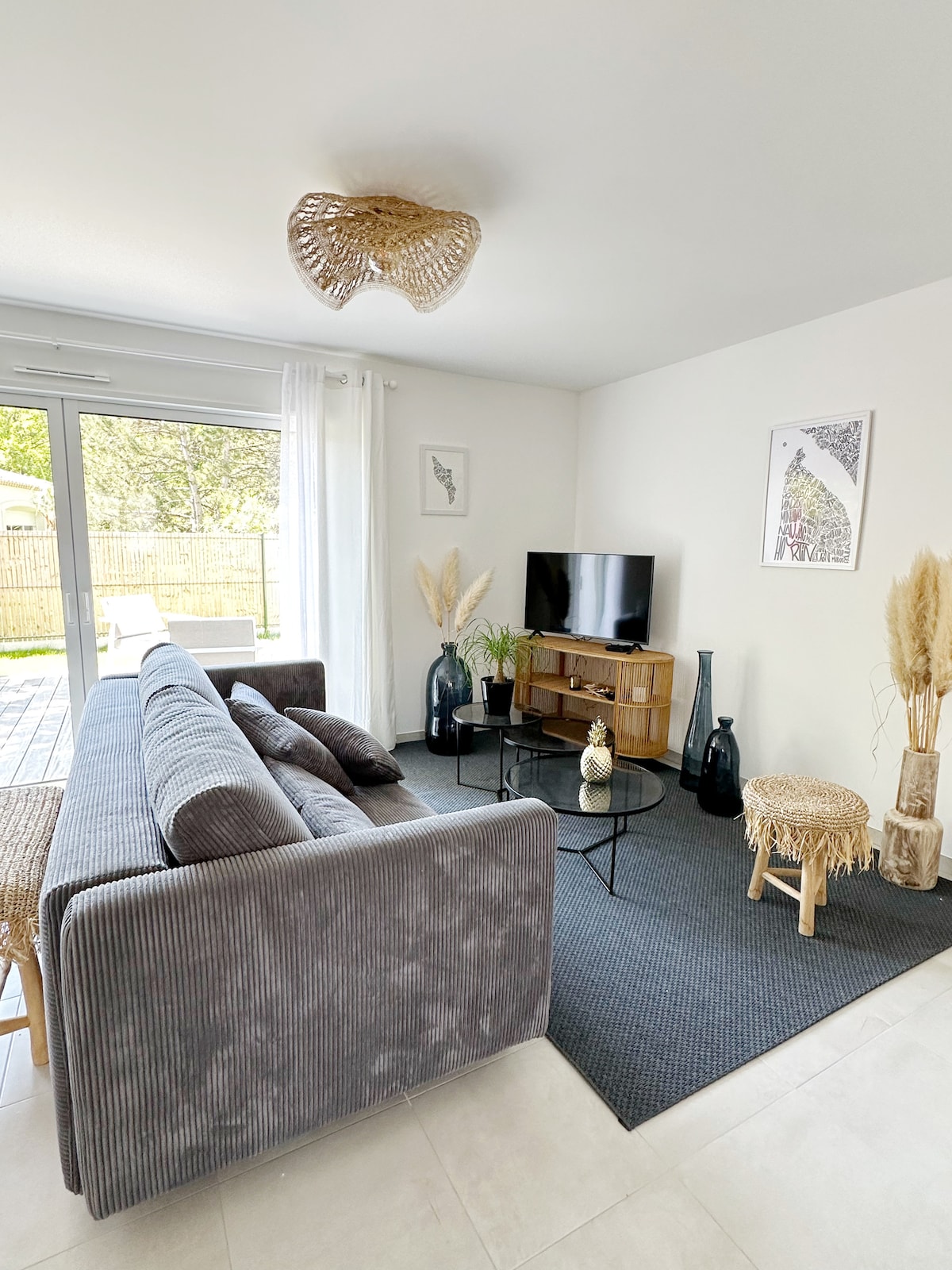A bright and airy living space features a comfortable, plush sofa with decorative pillows. A low coffee table sits in front, surrounded by minimalist decor including tall vases and artistic prints. Natural light streams in through large glass doors, illuminating the inviting atmosphere.