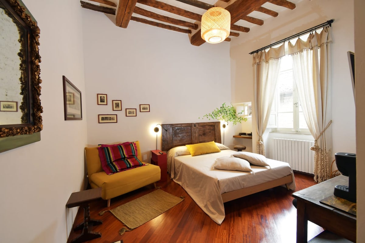 A spacious room features a wooden beam ceiling, complemented by natural light filtering through large curtains. A cozy bed is accompanied by decorative pillows, while a small sofa with colorful cushions offers additional seating. Framed artwork adorns the walls, enhancing the room's character.