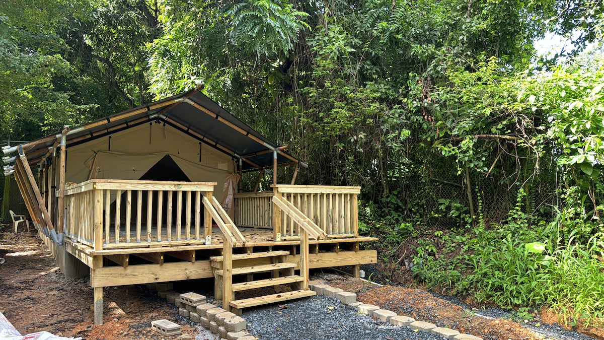 A spacious luxury tent is set on a wooden platform surrounded by lush greenery. Sturdy wooden steps lead up to the entrance, which is partially covered by a canopy. The natural landscaping provides a serene, private atmosphere.