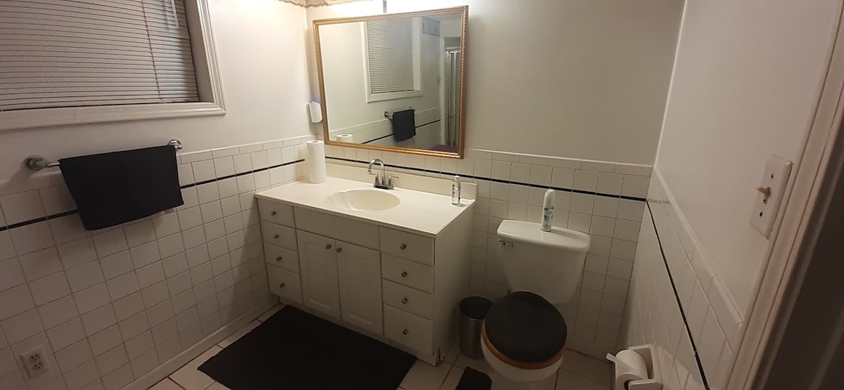The bathroom features white tiled walls and flooring, complemented by a large vanity with multiple drawers and a sink. A wall-mounted mirror with a wooden frame adds depth, while a shower is visible at the back. Towels are neatly hung on the rack.