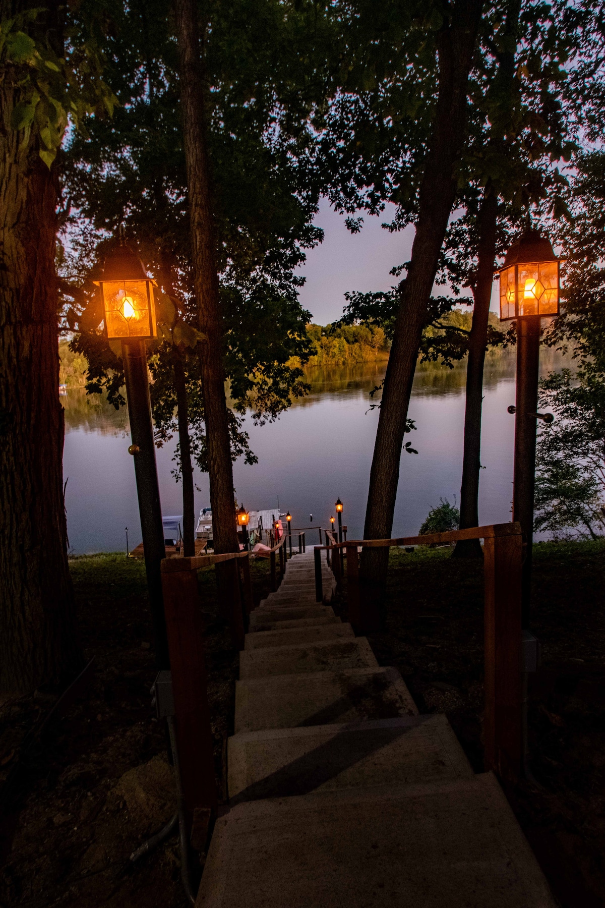 A stone staircase leads down to a serene lakeside, framed by trees and softly illuminated by lanterns. The water reflects the fading light of dusk, creating a calming atmosphere that invites leisurely strolls toward the private dock.